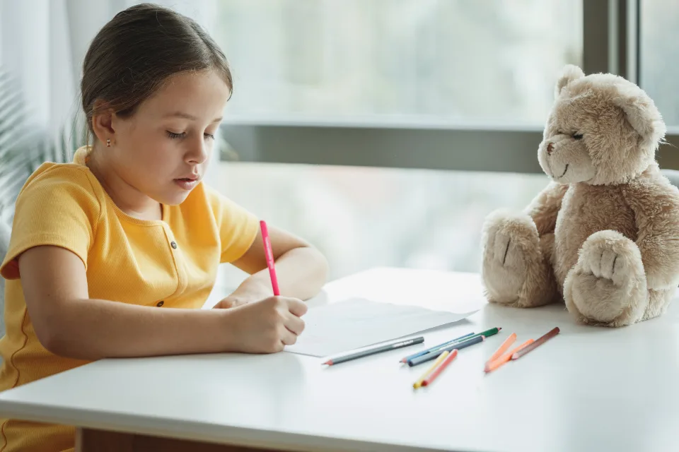 Child writing at a desk with pencils and a teddy bear
